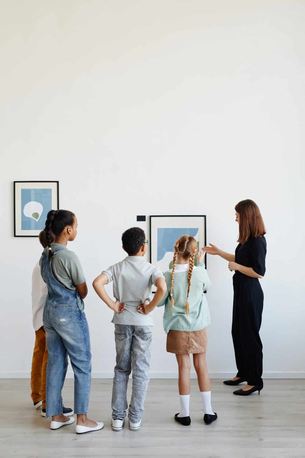 Vertical back view at diverse group of children listening to female tour guide while visiting modern art gallery